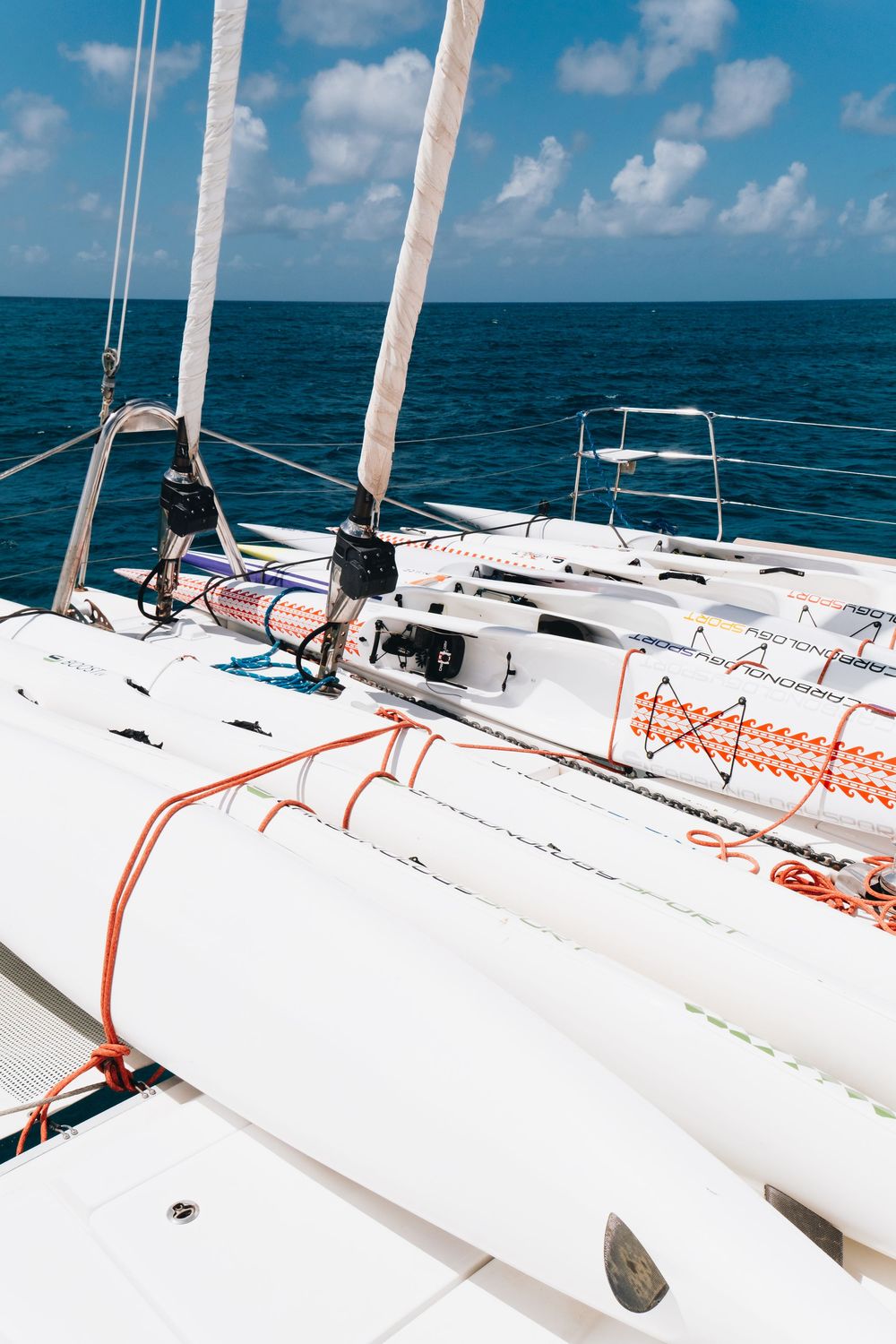 View from the deck of a sailing yacht showing white rigging, orange ropes and turquoise ocean waters under blue skies.