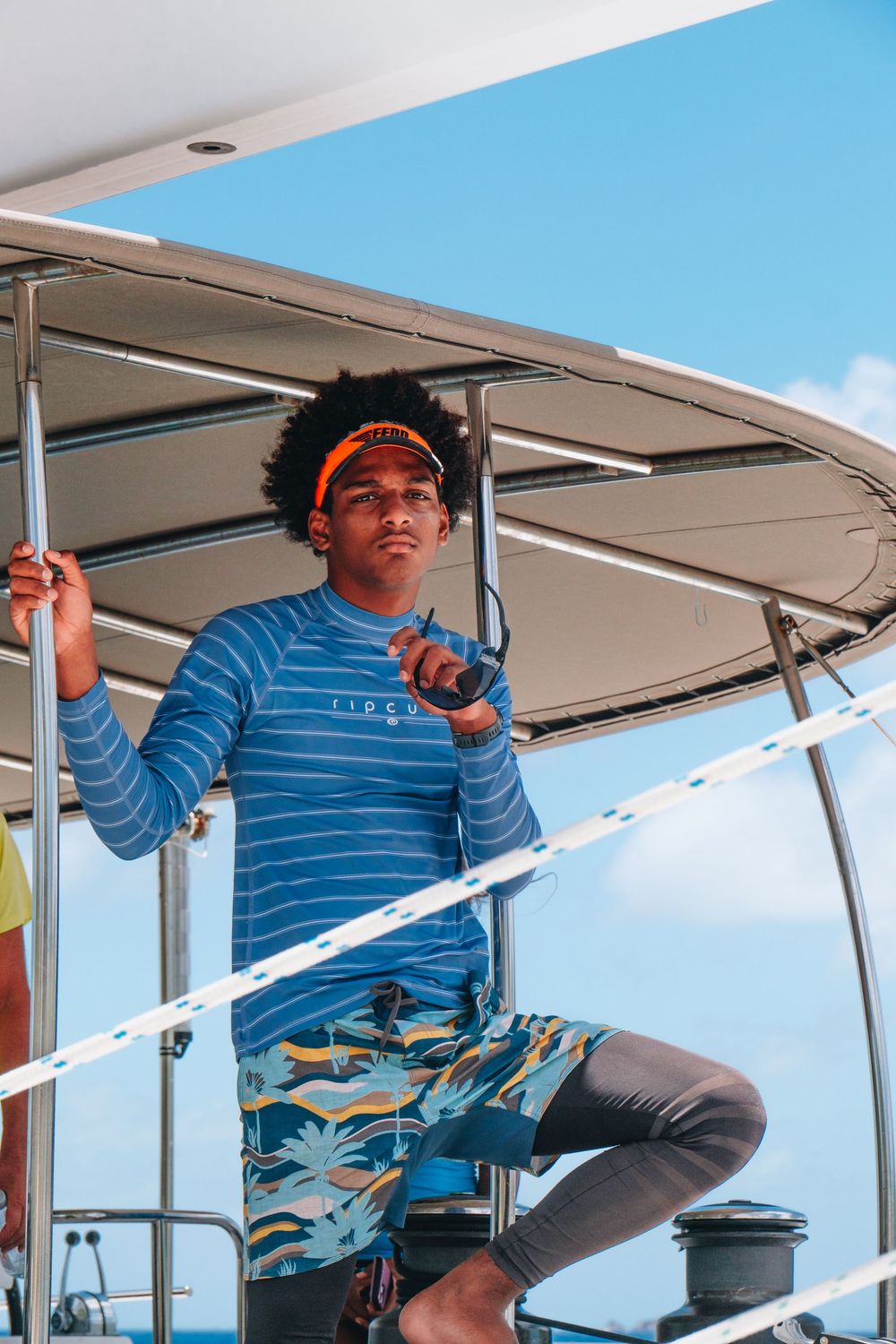 Person in blue striped shirt and floral shorts standing at boat helm against blue sky on sunny day.