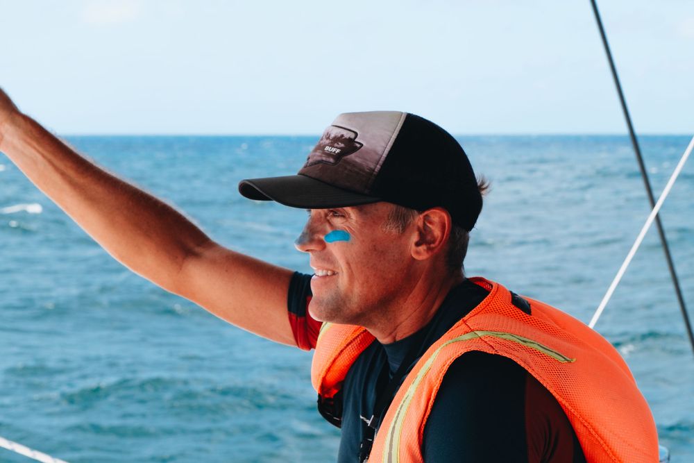 Boater wearing an orange life vest and baseball cap points while sailing on bright blue waters.