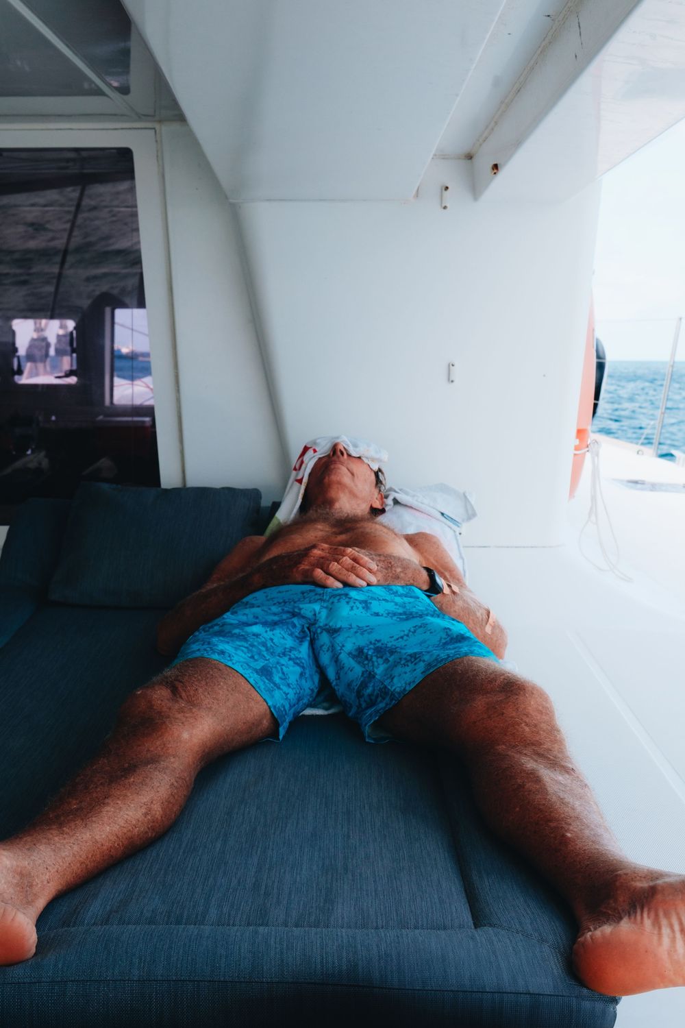 Person relaxing on a blue cushioned bench on a boat deck with ocean view visible through window.