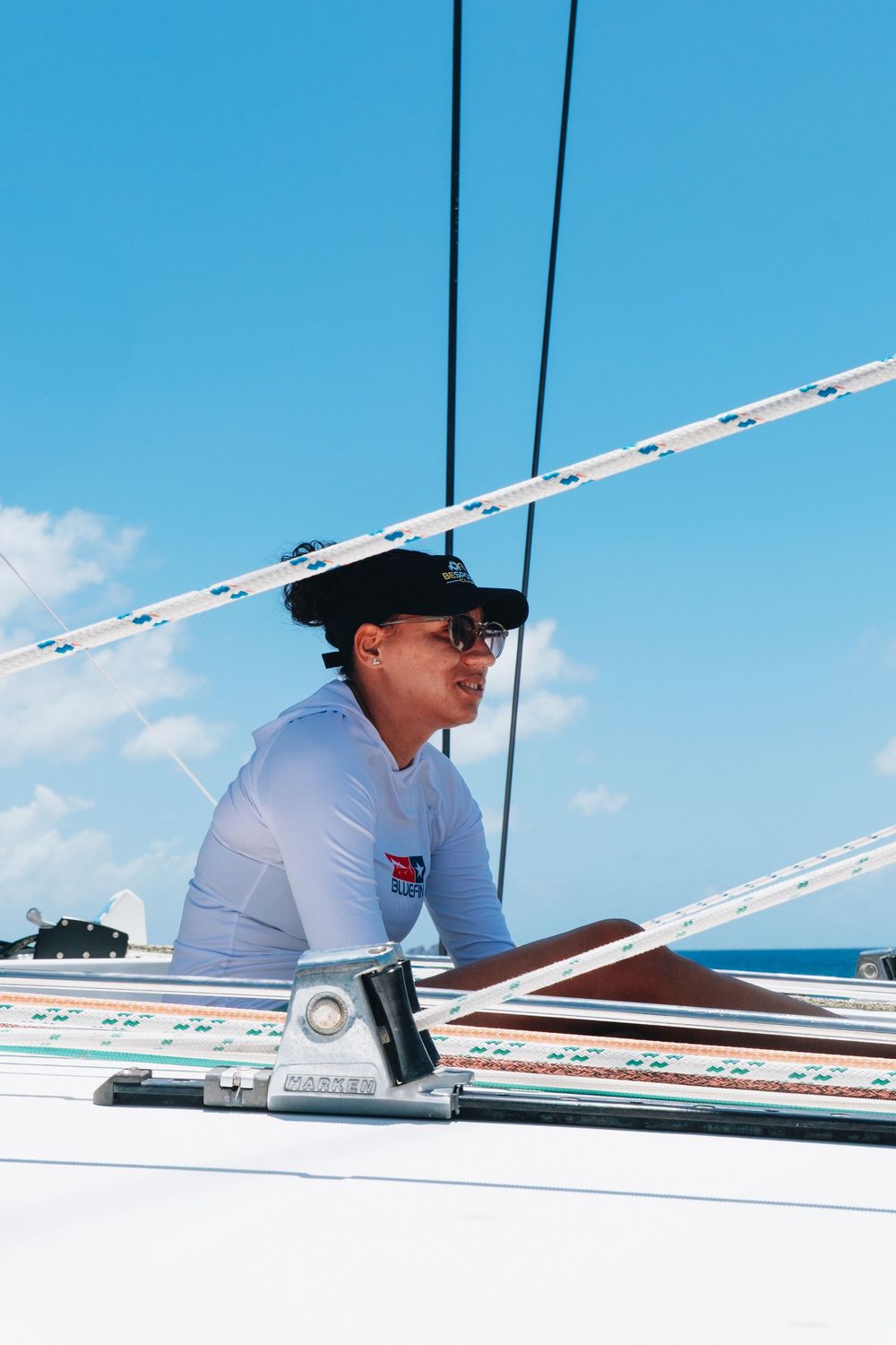 Person in white shirt and black hat sailing on a boat against bright blue sky with rigging visible.