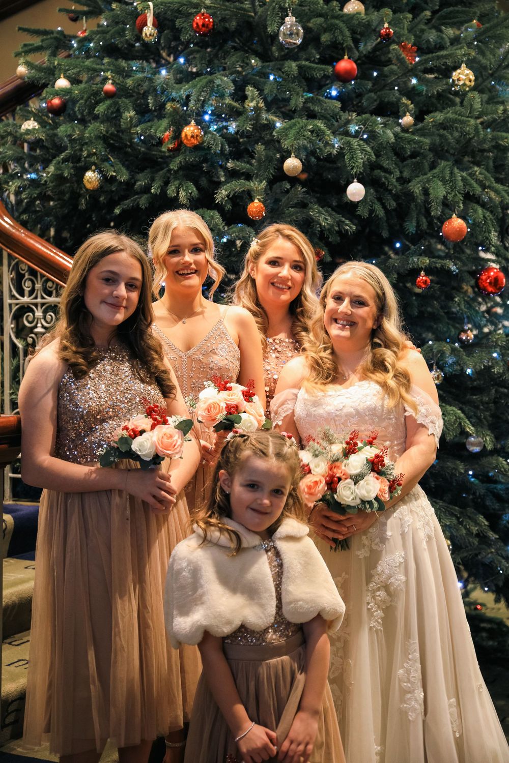A group in formal attire poses in front of a decorated Christmas tree with flowers and holiday ornaments.