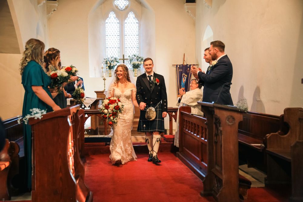 Newlyweds exit the church aisle together after ceremony in traditional church with stained glass windows and wooden pews.