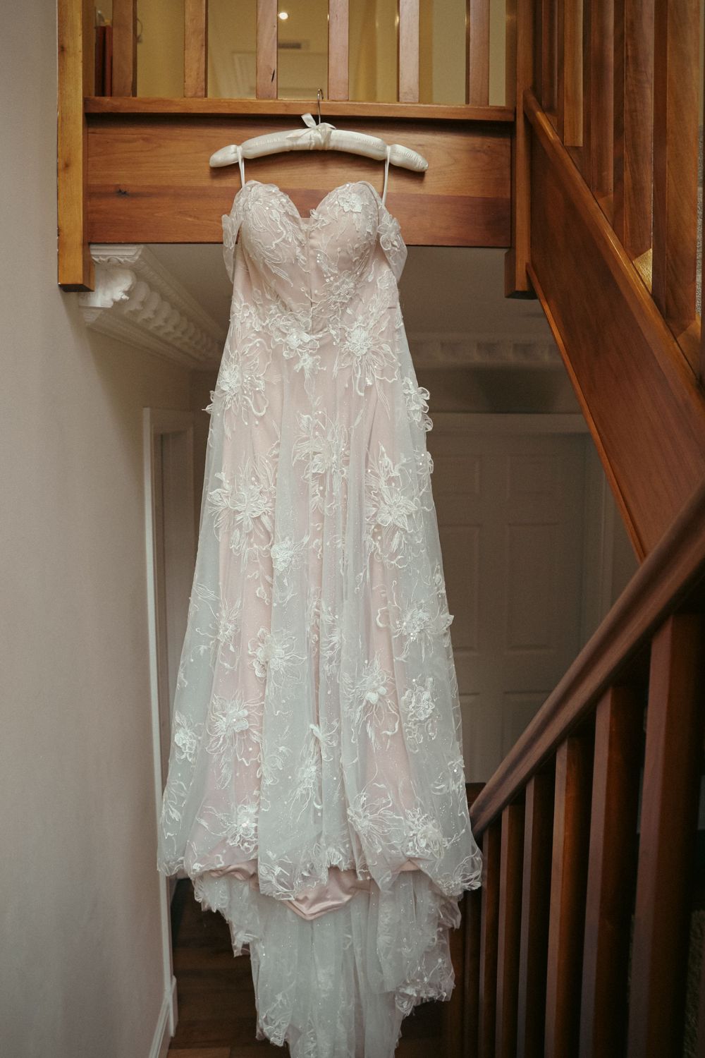 Beautiful white lace wedding dress with floral appliques hanging on wooden hanger against staircase.