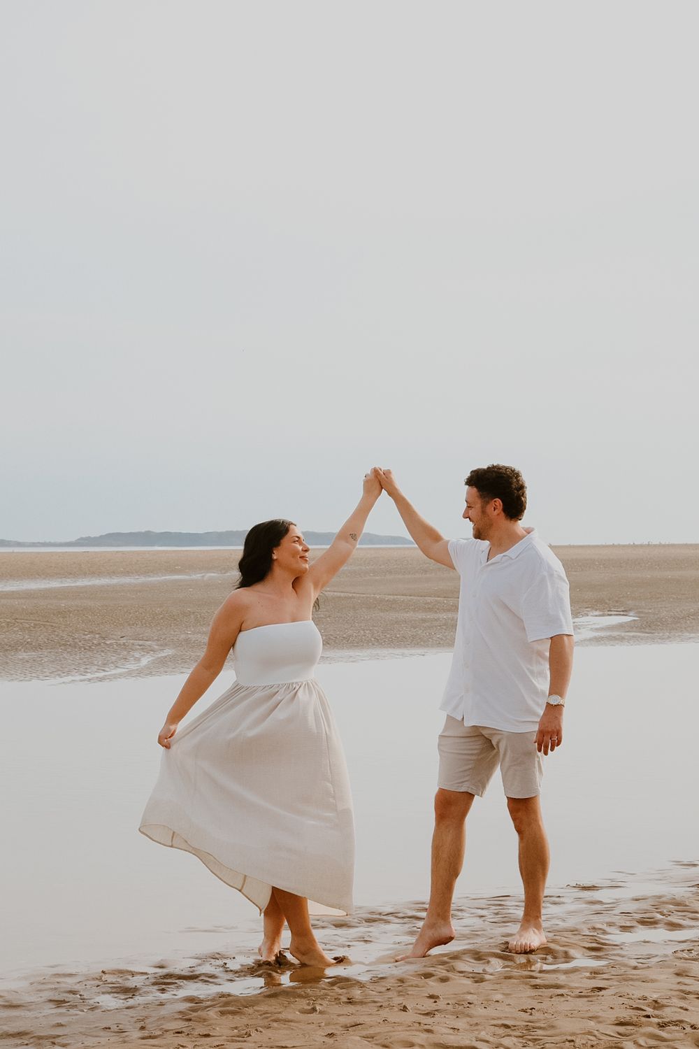 Couple in white outfits dancing playfully along a misty beach at sunset, creating a romantic scene.