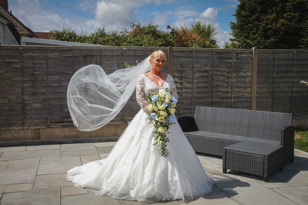 A flowing white wedding dress and veil catch the breeze in an outdoor garden patio setting with grey furniture.