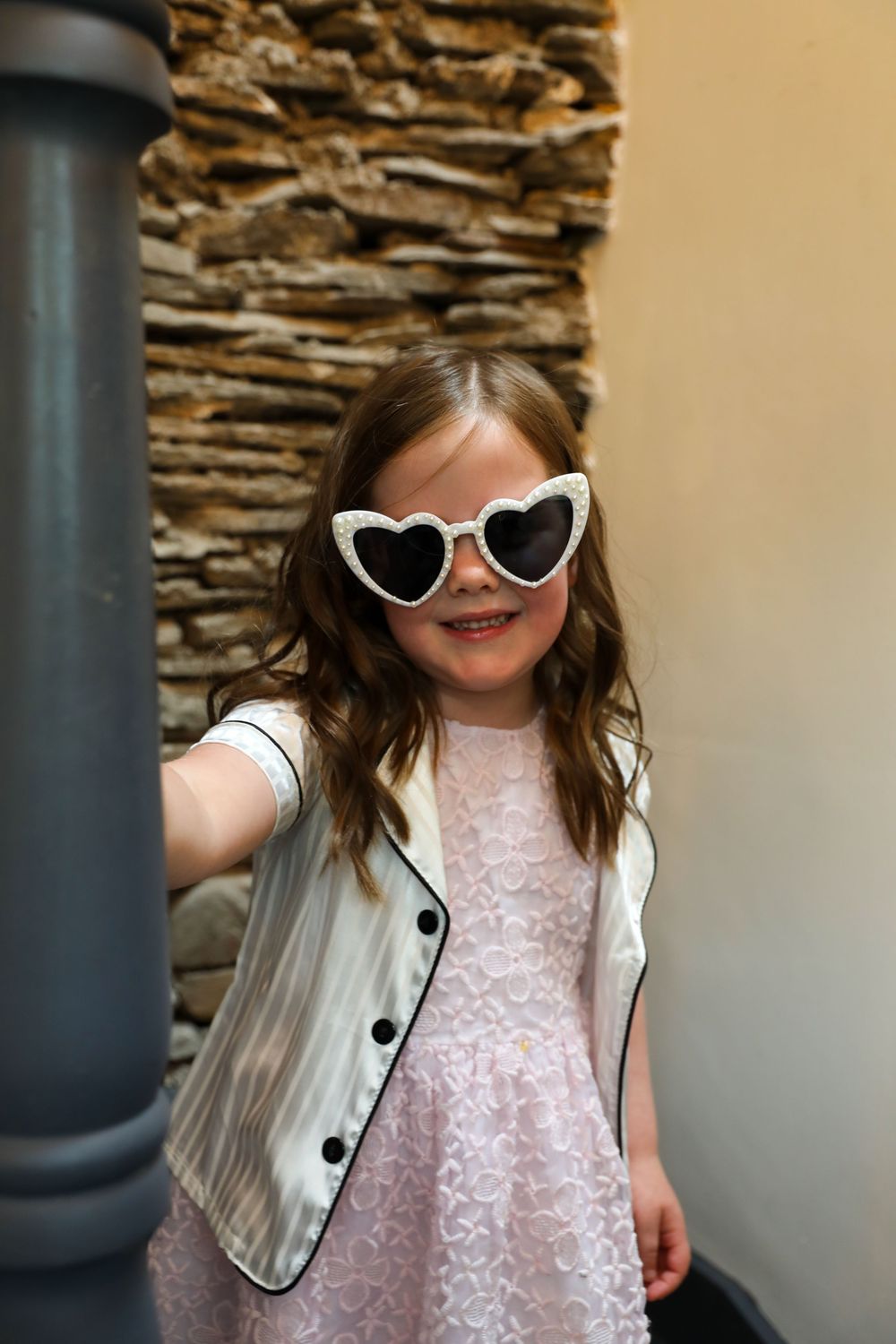 Child in a pink lace dress and white vest wearing heart-shaped sunglasses poses against a stone wall.
