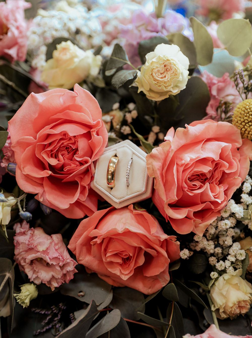 Beautiful coral pink roses surrounding a hexagonal ring box with wedding rings in a romantic floral arrangement.