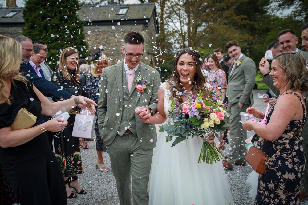 A joyful wedding couple walks through flying confetti surrounded by guests at an outdoor venue.