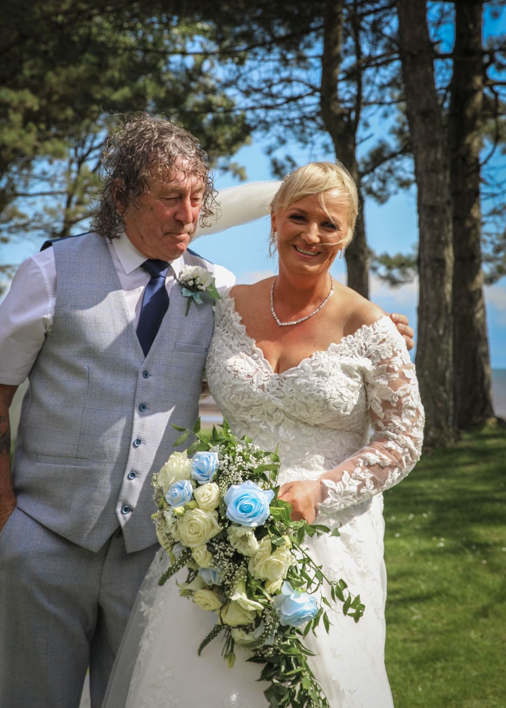 A couple in wedding attire pose outdoors with a beautiful blue and white floral bouquet on a sunny day with pine trees behind.
