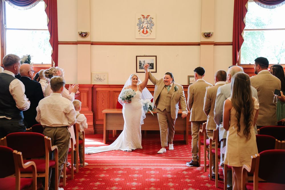 Bride and groom walk down the aisle at a wedding ceremony in an elegant wood-paneled hall with seated guests.