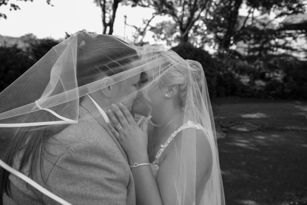 Black and white photo of two brides sharing a romantic kiss under a wedding veil outdoors.