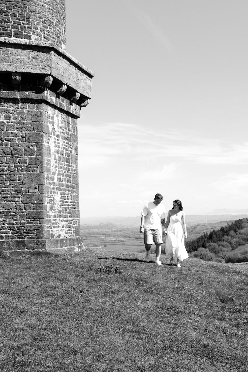 Black and white photo of a couple holding hands walking near a historic stone tower with scenic countryside views.