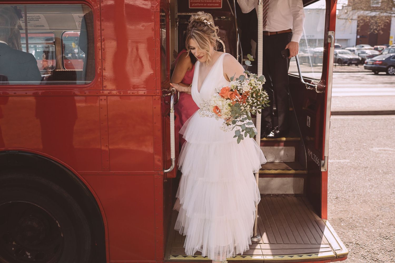 A bride in a white wedding dress holding a bouquet boards a vintage red double-decker bus.