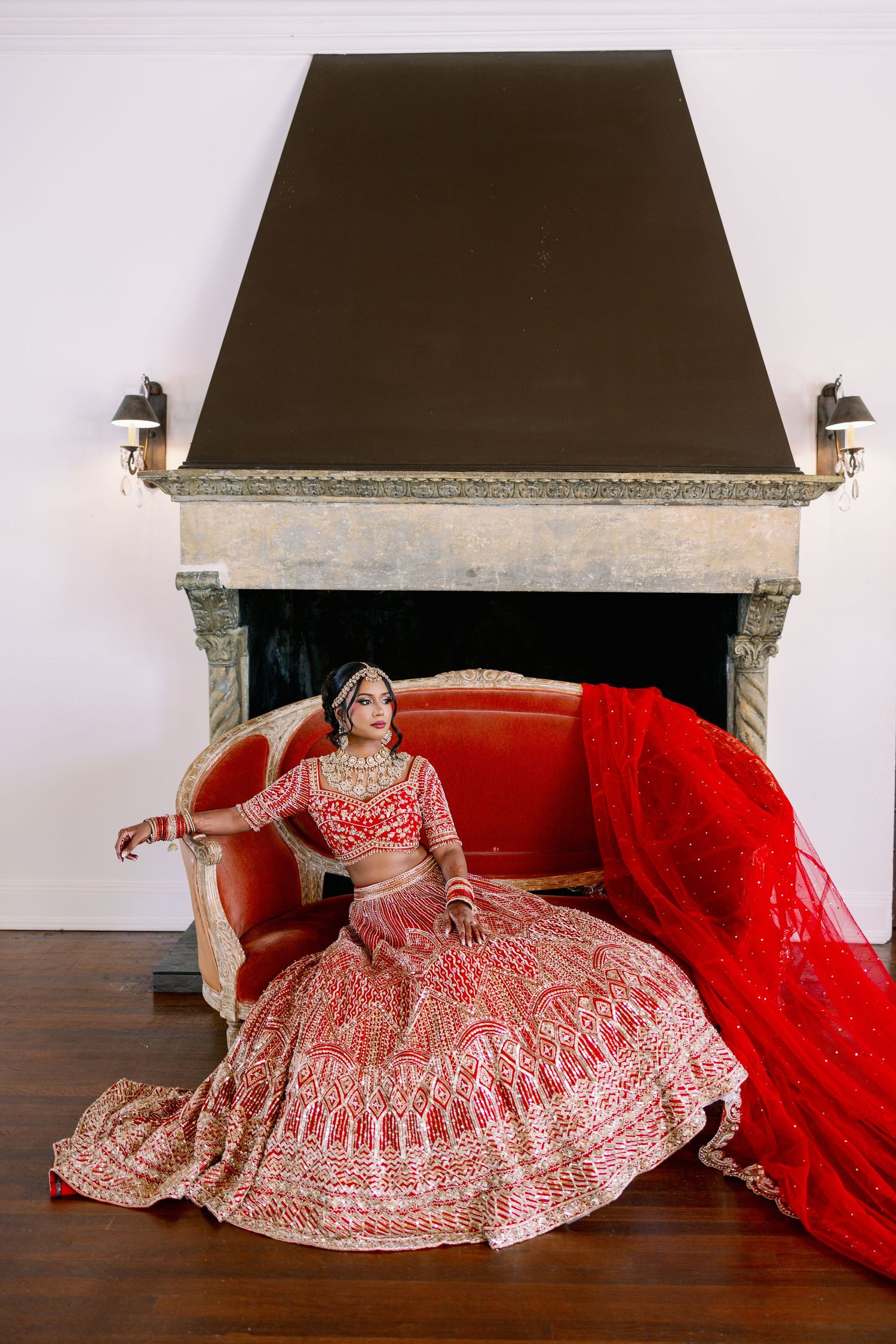 Traditional Indian bride in red and gold embroidered lehenga poses elegantly on red couch near fireplace.