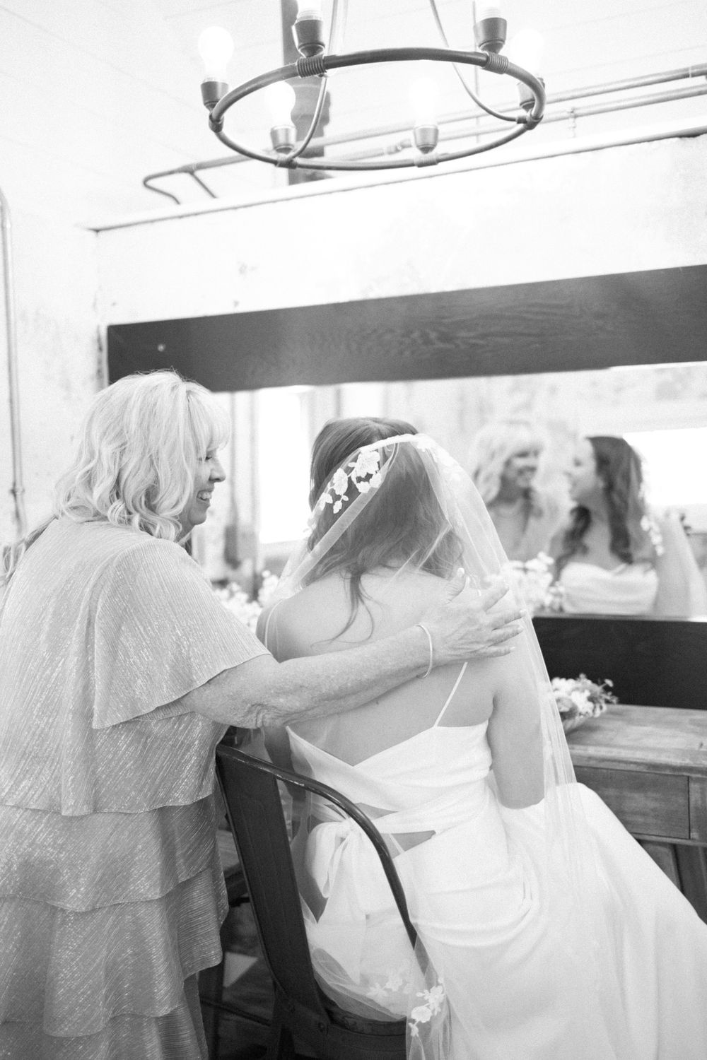 A bride in a white wedding dress sits at a vanity mirror while someone helps with final preparations in vernon nj.