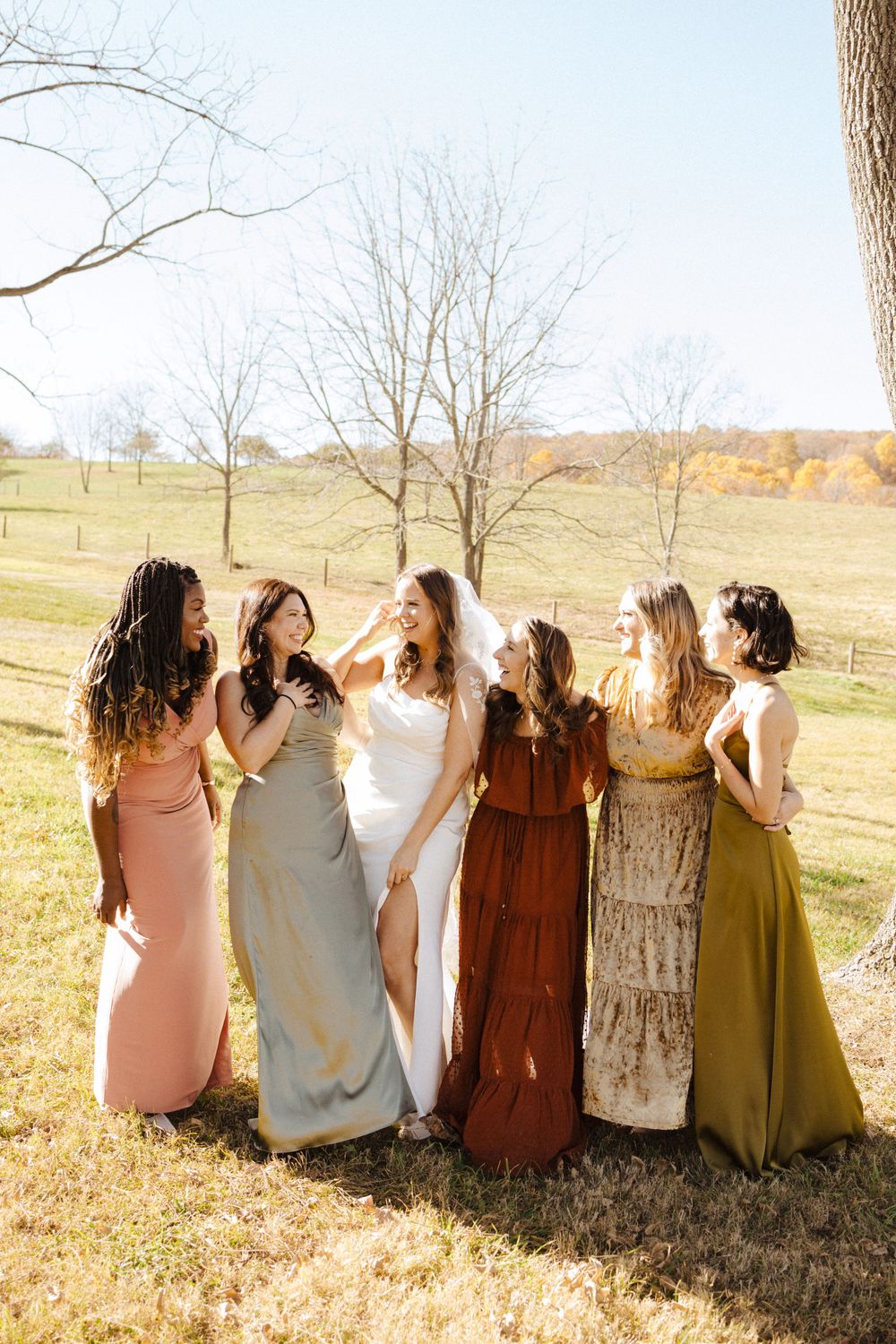 A group in earth-toned dresses stand together in a meadow during golden hour with autumn trees in the background  in vernon nj.