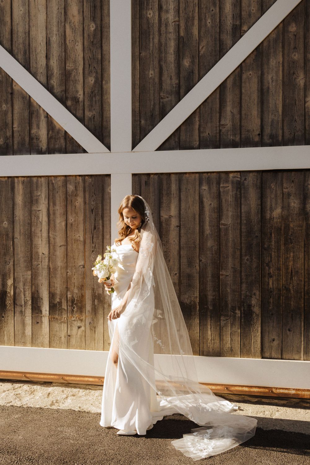Bride in elegant white gown poses against rustic barn wall with white trim.