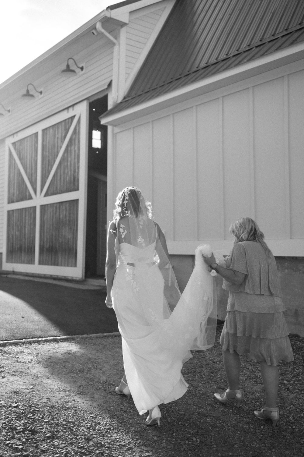 A bride in a flowing white gown walks toward a rustic barn building while her dress is held by attendants behind her in vernon nj.