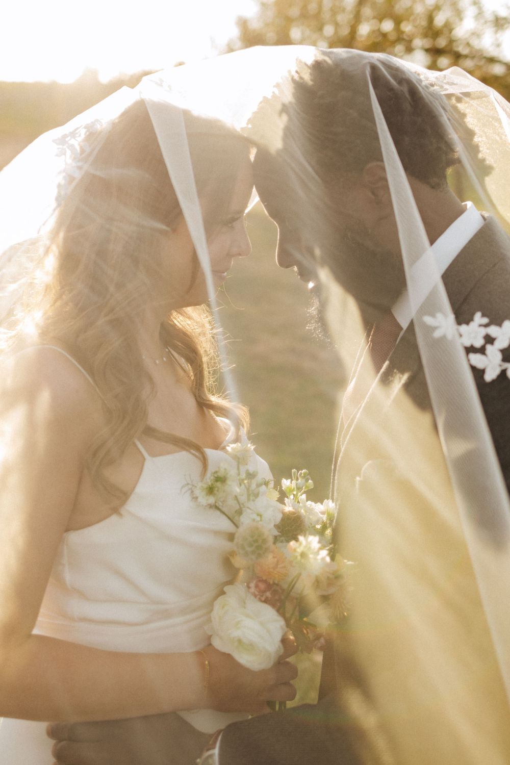 A bride and groom share an intimate moment under her veil during sunset with a bouquet of white flowers in frenchtown nj.