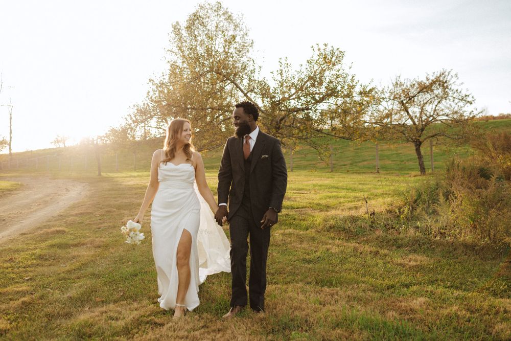 Couple poses in sunlit field with autumn trees and rolling hills in background.