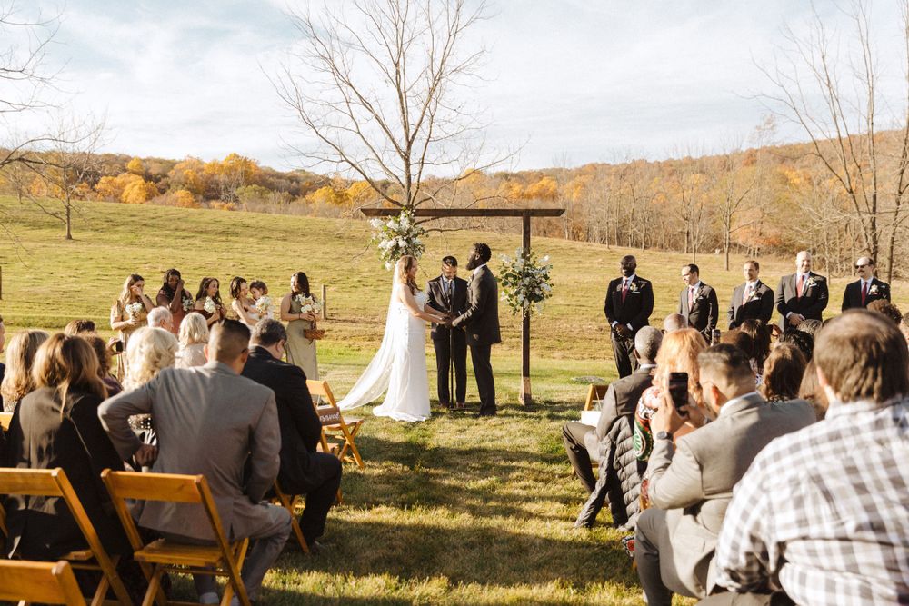 Outdoor autumn wedding ceremony with guests seated on wooden chairs facing an altar.
