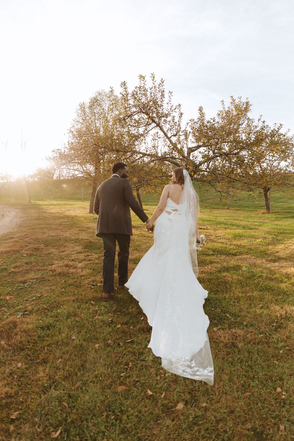 Newlyweds walk hand in hand through an orchard at sunset with flowing wedding dress.