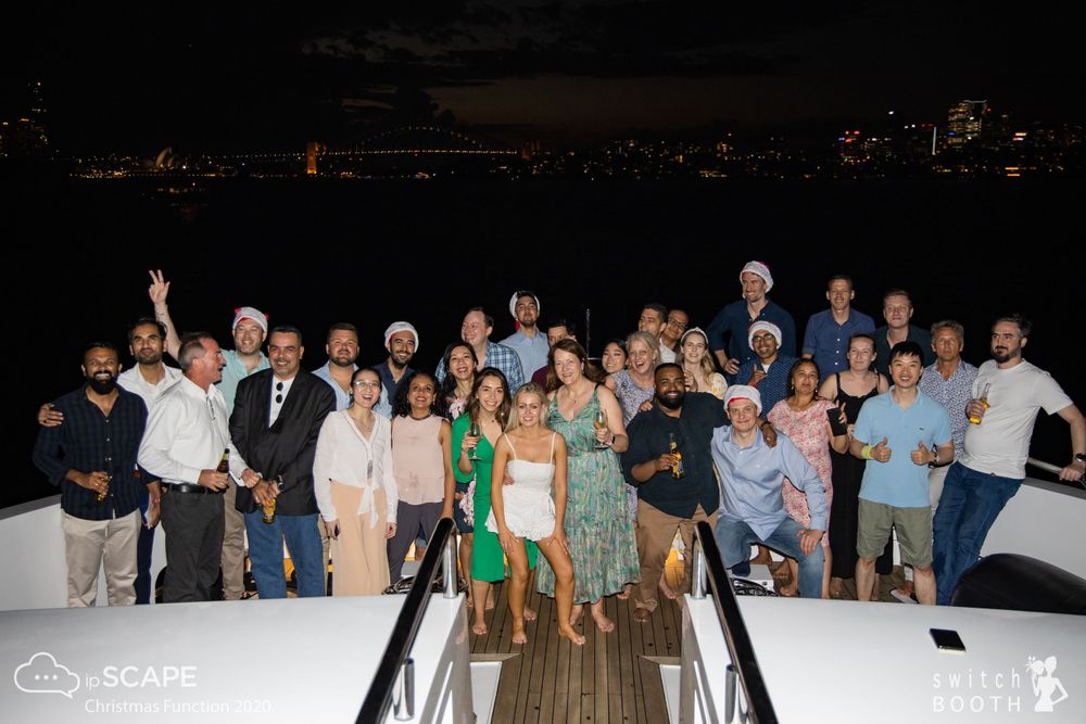 Sydney Event Photography - Large group poses together for a nighttime photo on the deck of a boat with city lights in background.