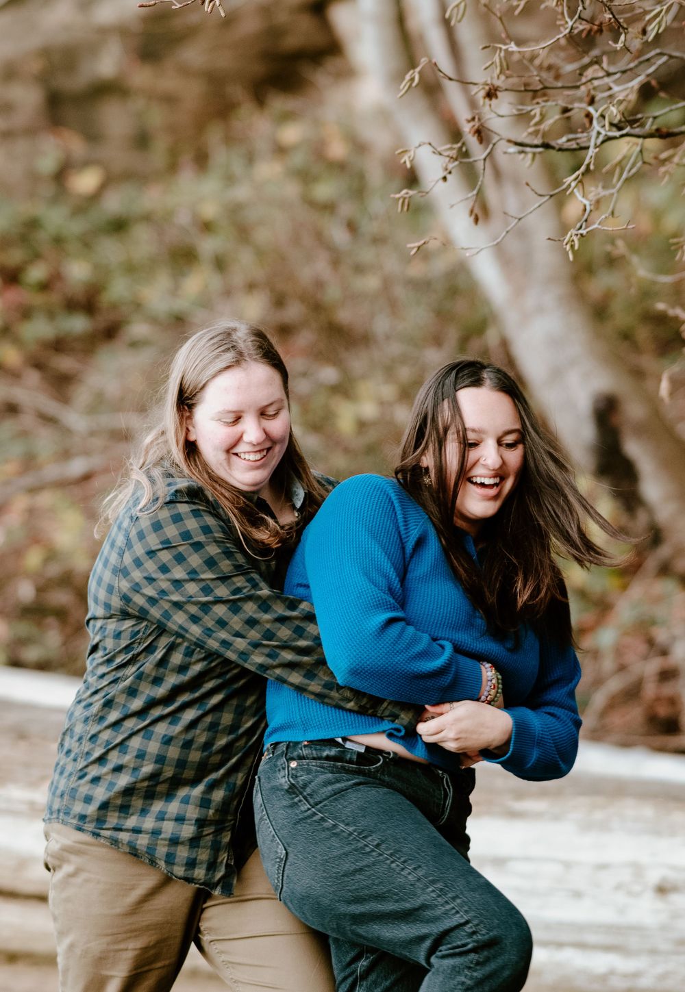 Two people in casual outdoor wear laughing and embracing by a creek.