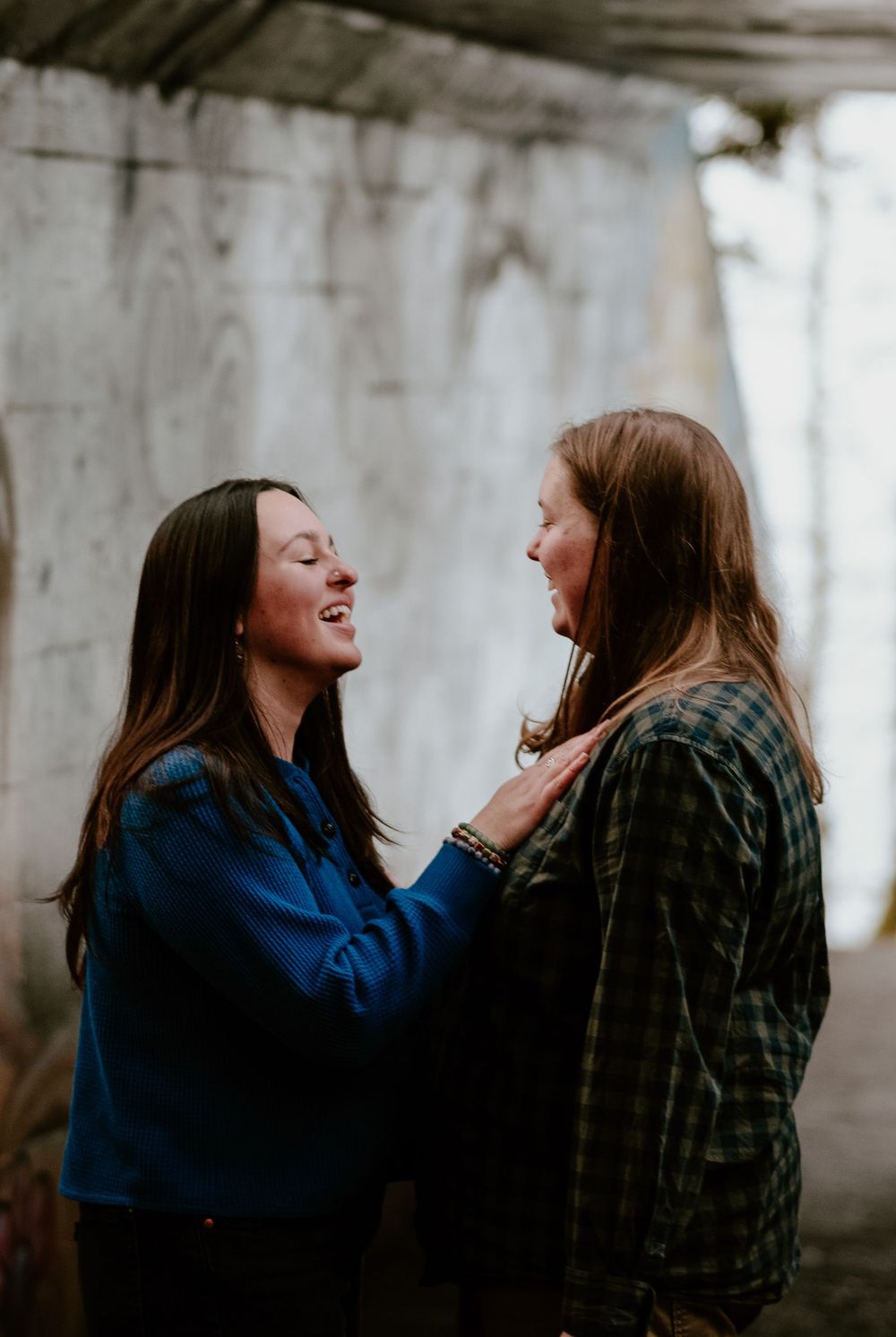 Two people laughing and embracing in front of concrete wall outdoors.