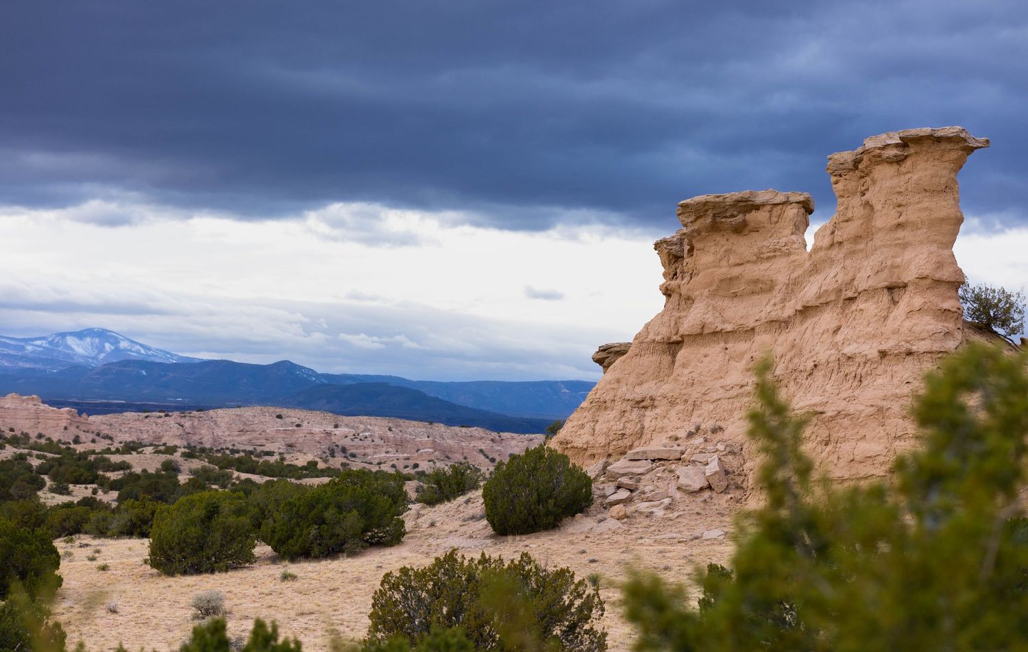Dramatic rock formations in a desert landscape with stormy skies and mountains in the distance.
