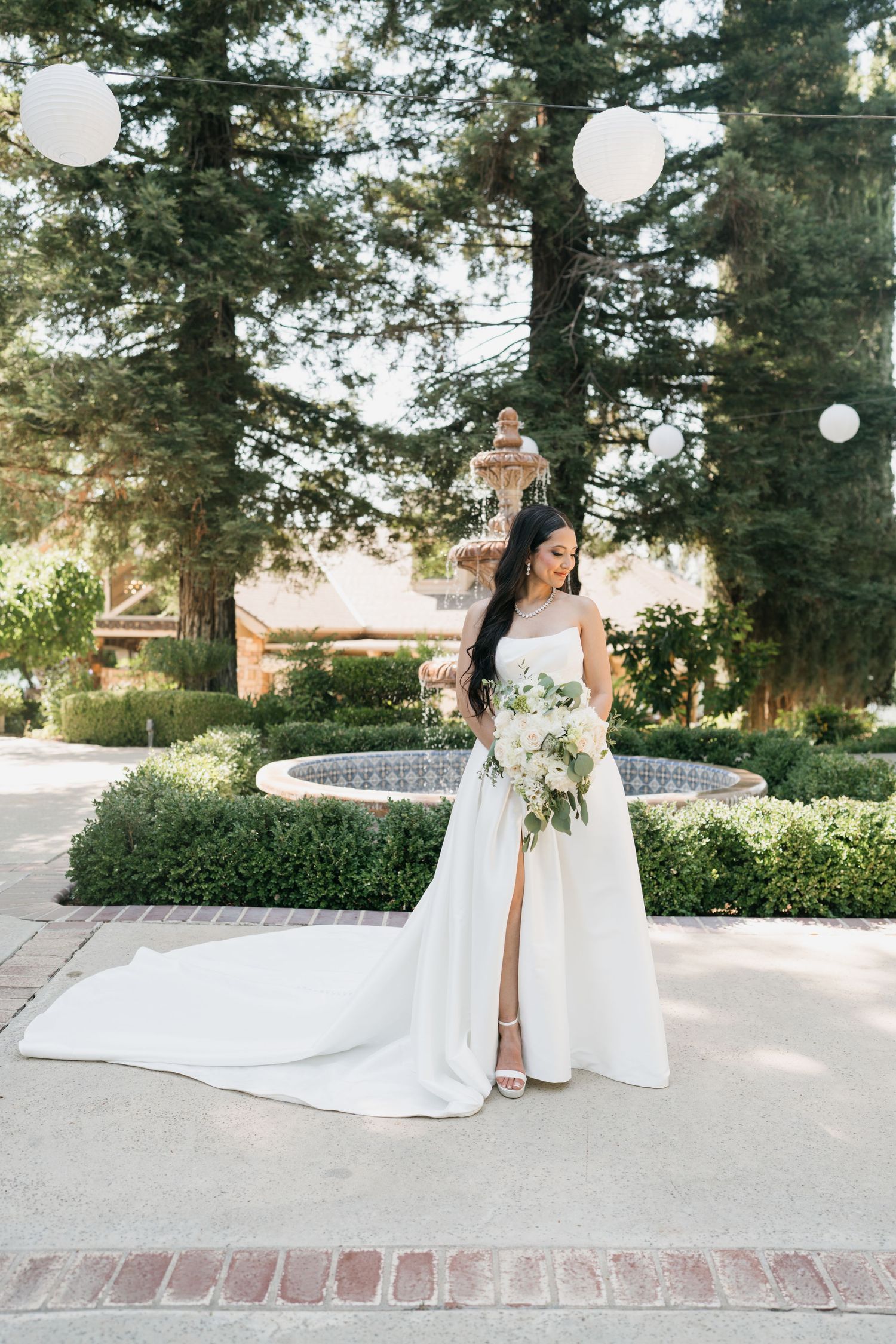 A bride in a white lace gown holds a classic white bouquet against a neutral background.