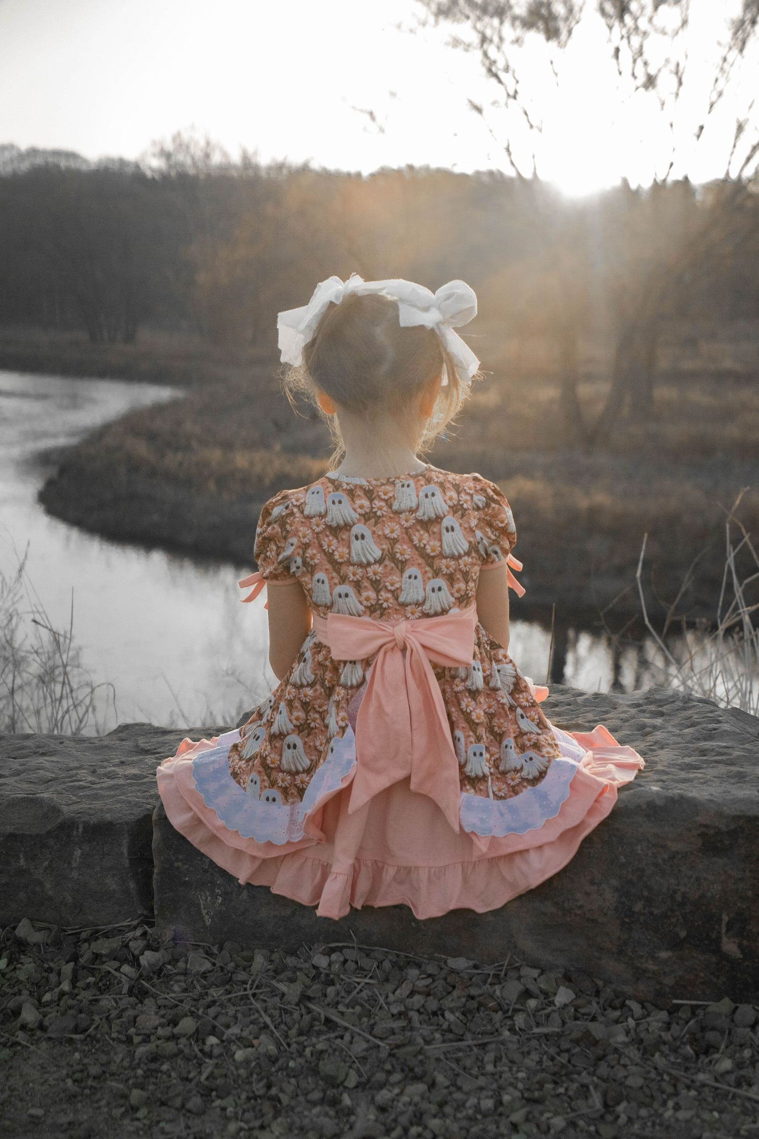 Young girl in ghost-print dress with pink bow sits on stone wall by river at golden hour.