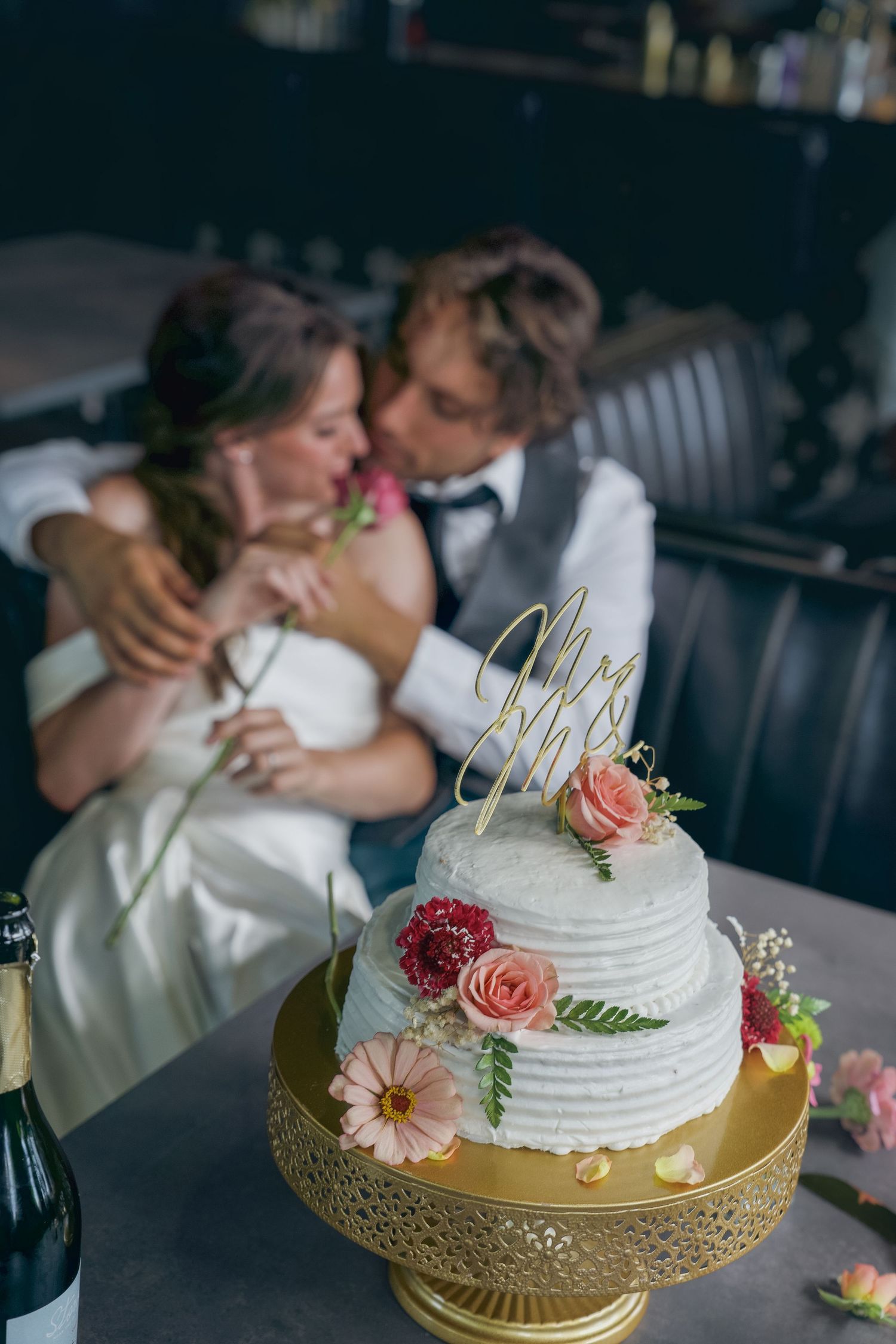 Two-tier white wedding cake with floral decorations and Mr topper on gold stand, with bride and groom kissing in background.