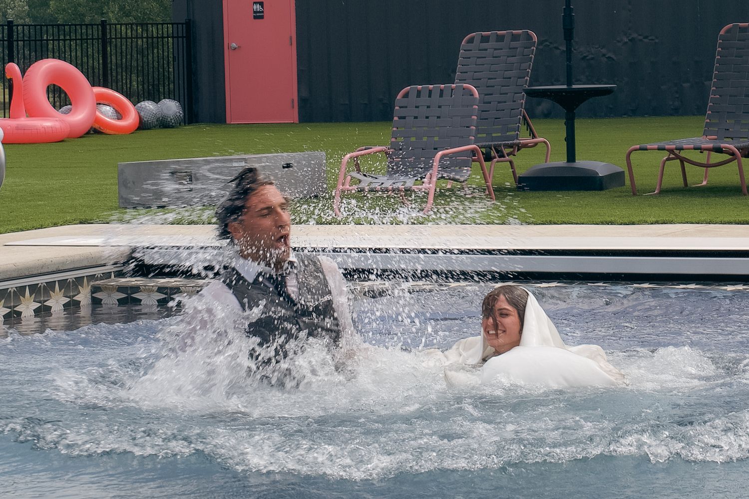 Two people splashing in a backyard pool with lawn chairs and pink flamingo floats visible nearby.