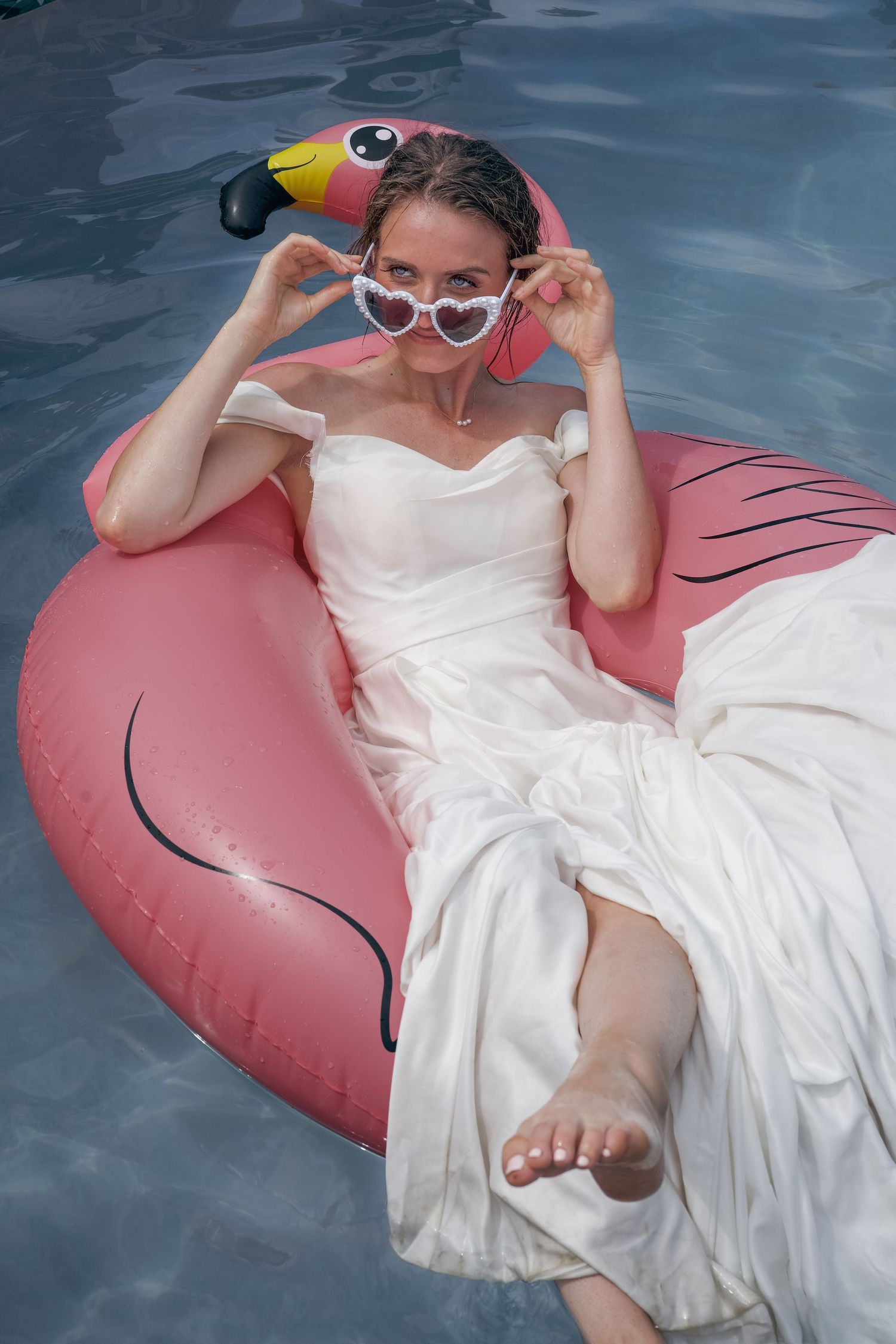 Bride in white wedding dress floating on pink flamingo pool float, wearing heart-shaped sunglasses in a swimming pool.