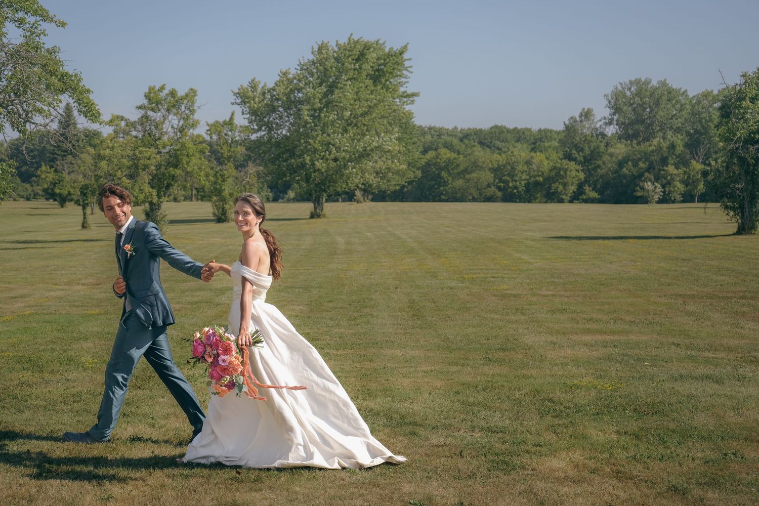 Bride in white gown holding pink bouquet walks hand-in-hand with groom in blue suit across lush green lawn.