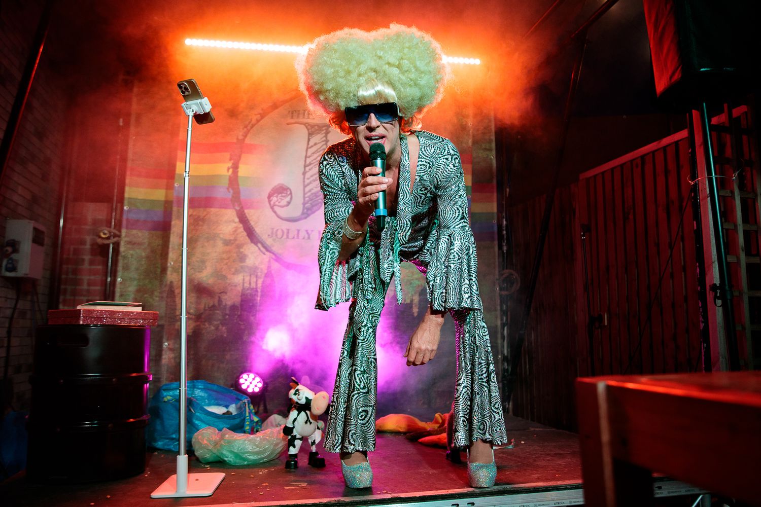 Drag performer in green sequin outfit and afro wig sings on a colorful, neon-lit stage.