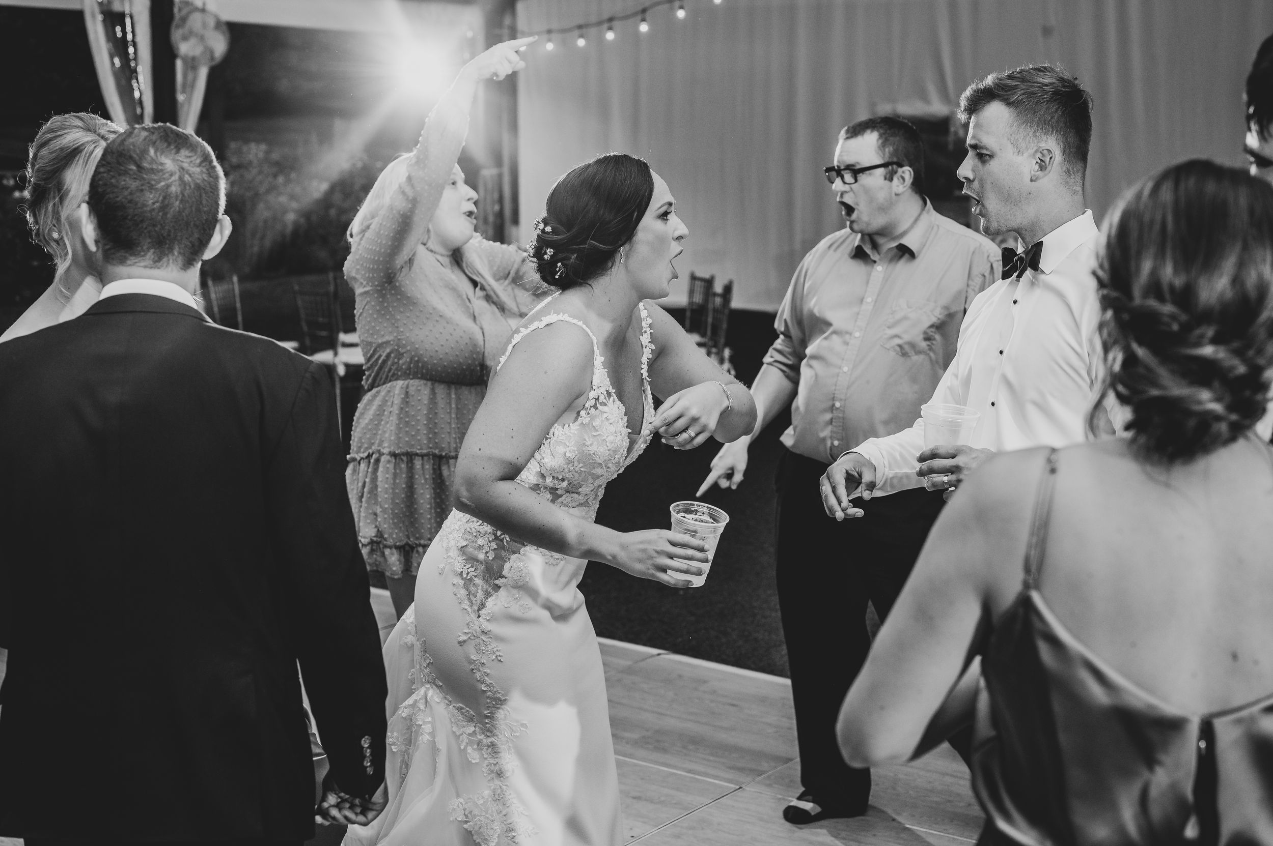 Black and white wedding photo of bride smiling at couple dancing in foreground, floral arch backdrop.
