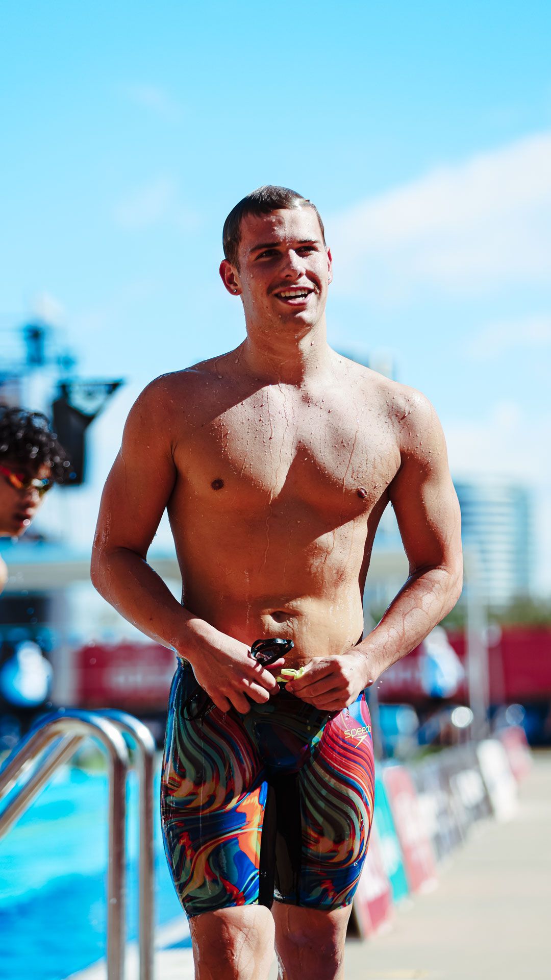 Smiling athletic young man in colorful swim trunks standing poolside at an outdoor swimming competition.