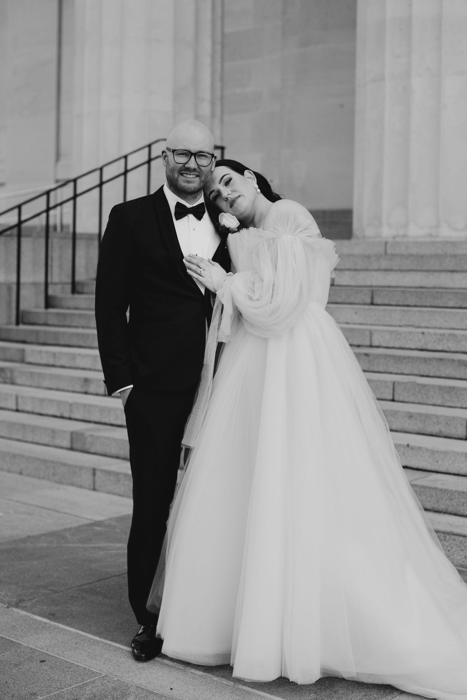 Bride in flowing white gown leaning on groom in black tuxedo on stone steps in black and white.