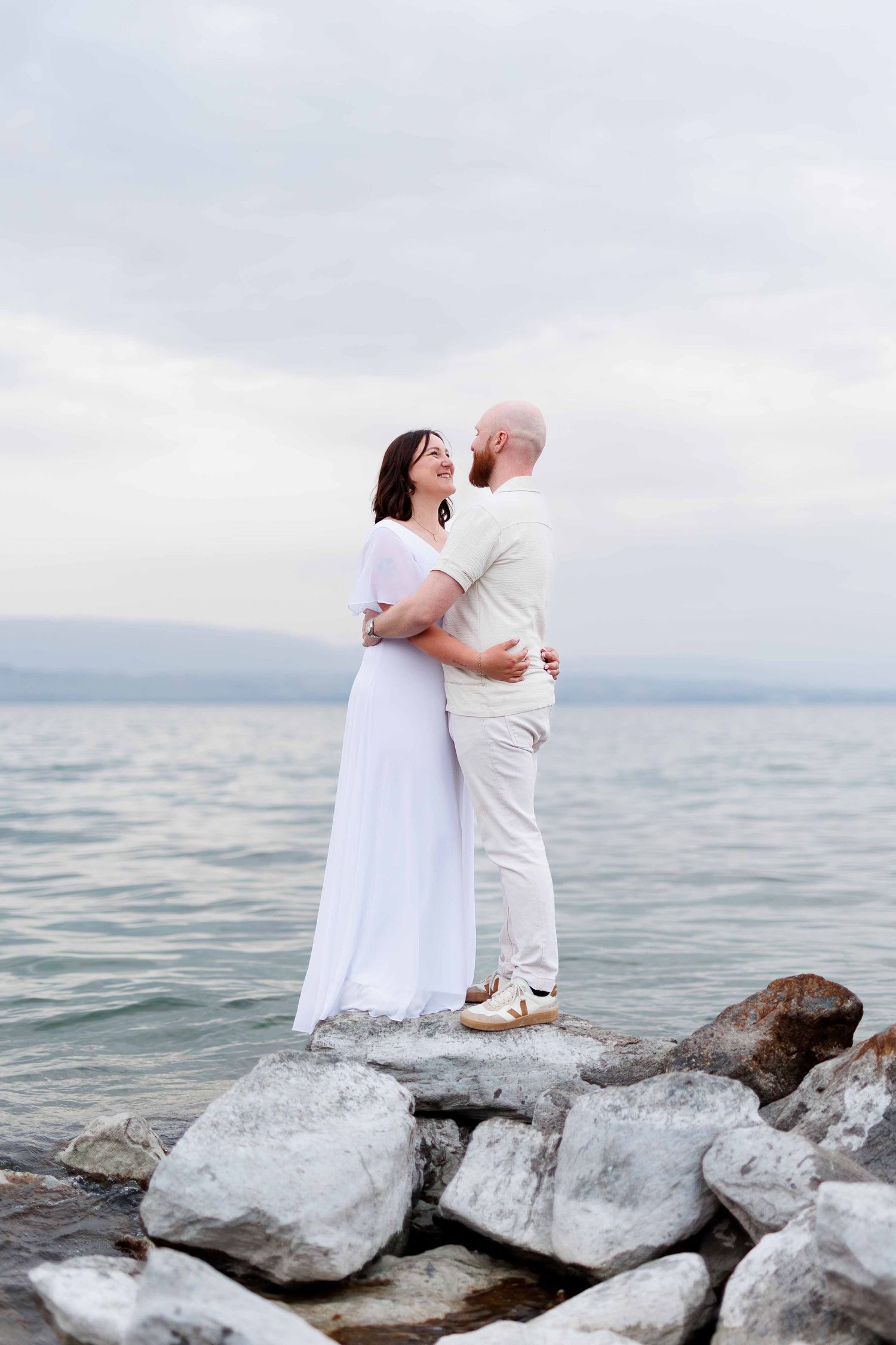 Couple embracing on rocky shoreline, woman in white dress, man in beige outfit, calm sea and cloudy sky backdrop.
