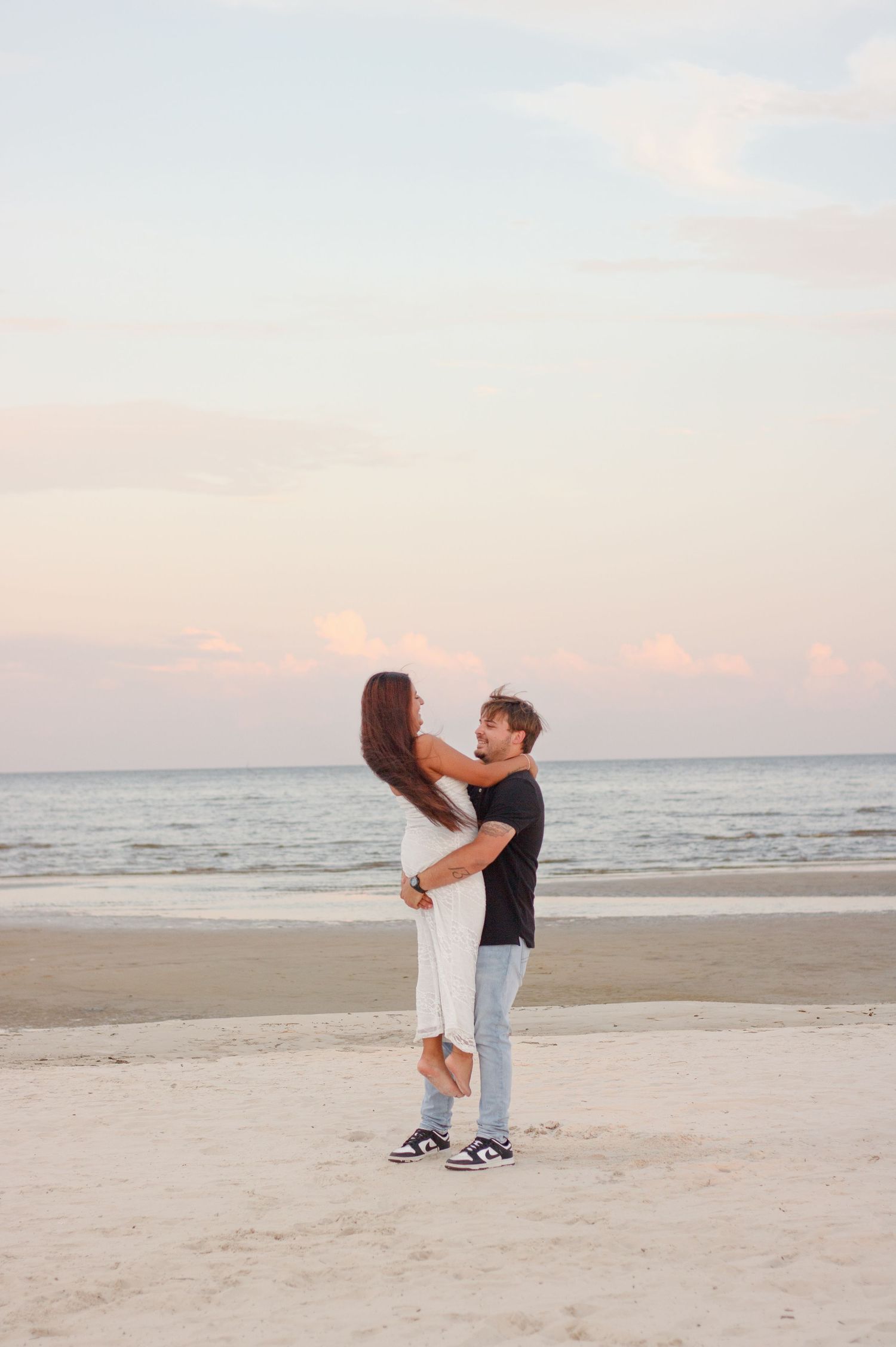 A couple embraces on a sandy beach as the woman is lifted in a hug under a soft pink sunset sky.