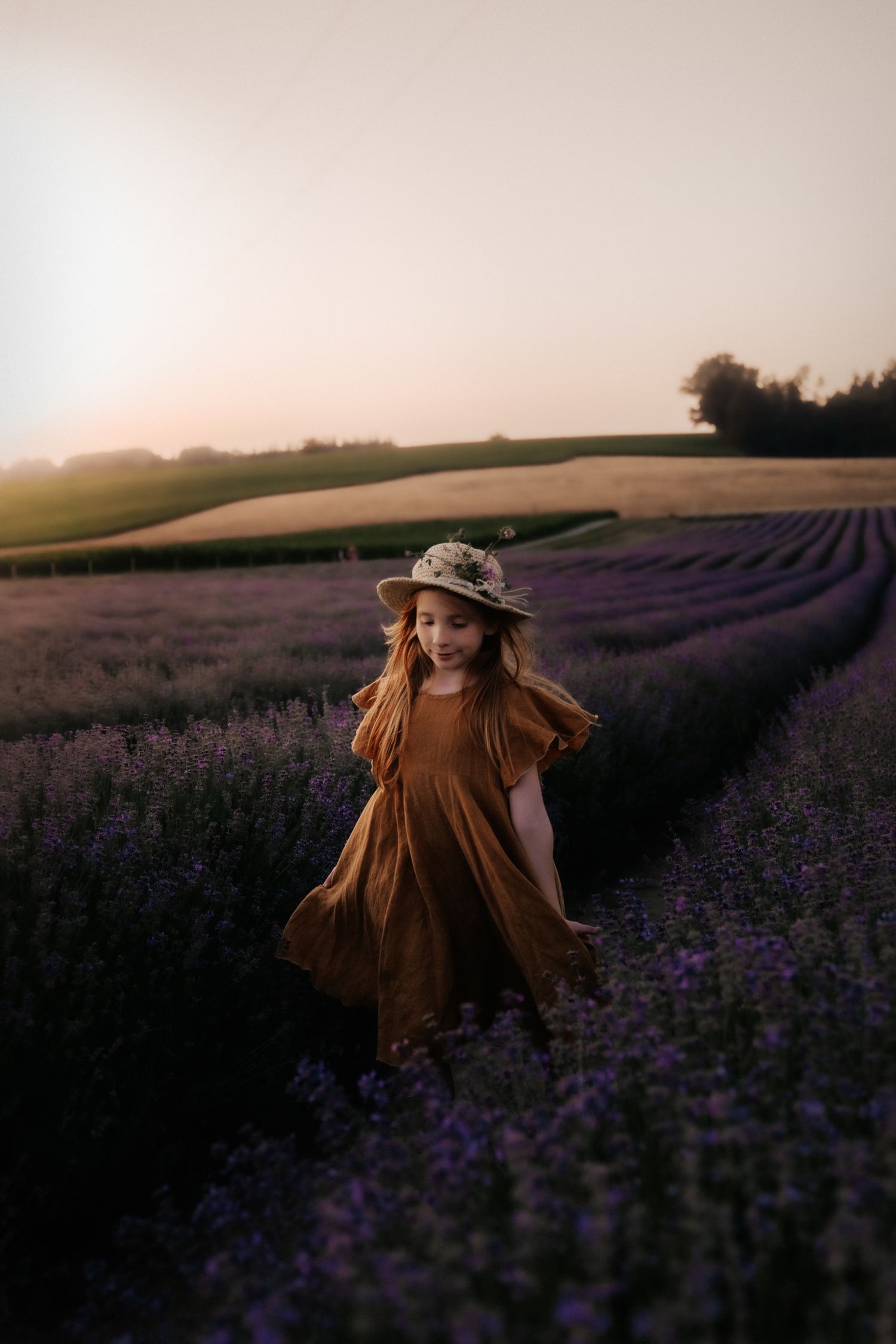 Young girl in brown dress and floral hat runs through purple lavender fields at golden sunset.