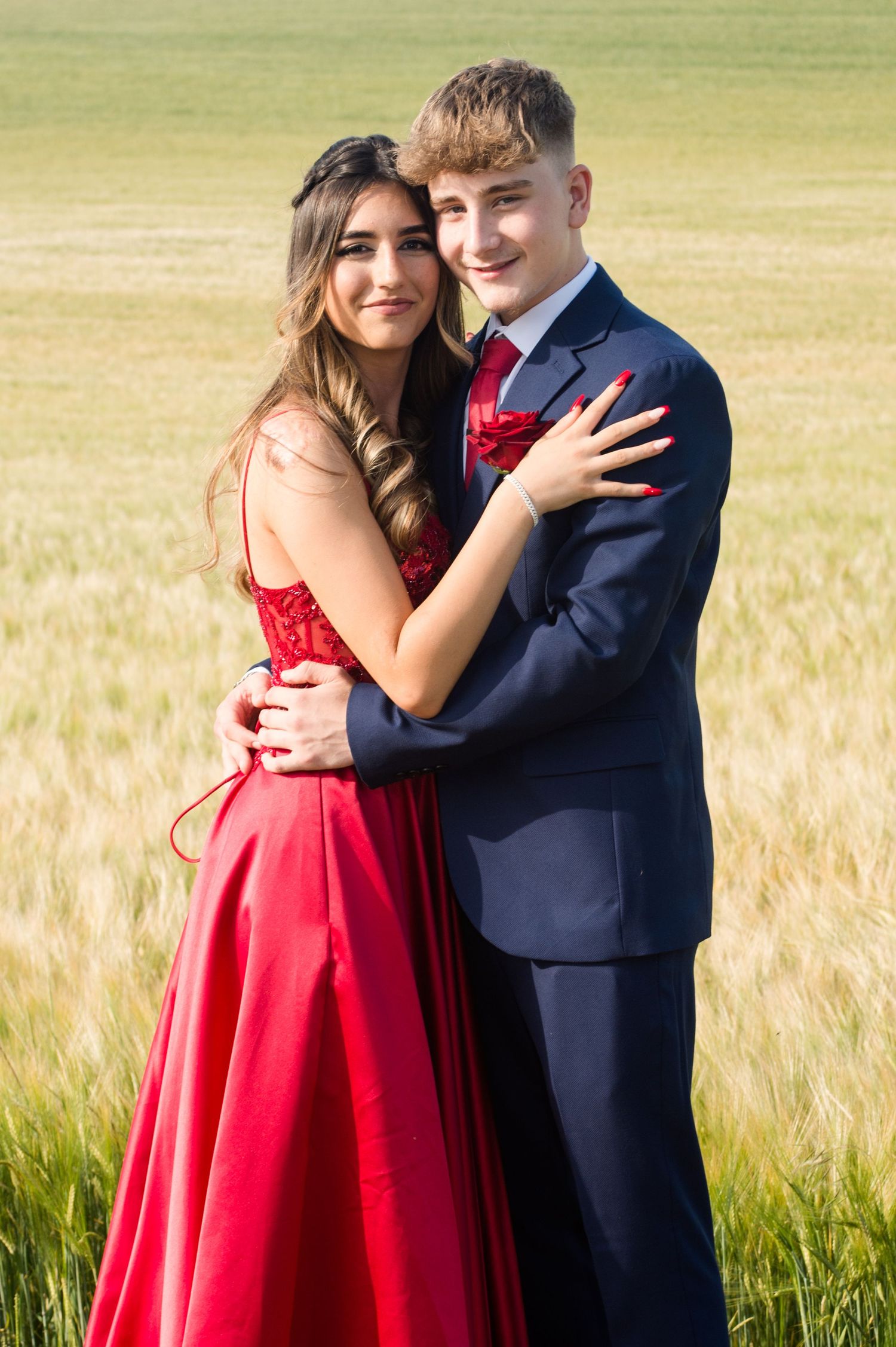 A couple stands in a field, embracing. The woman wears a red dress and the man a navy suit. Both smile warmly under a sunny sky, suggesting