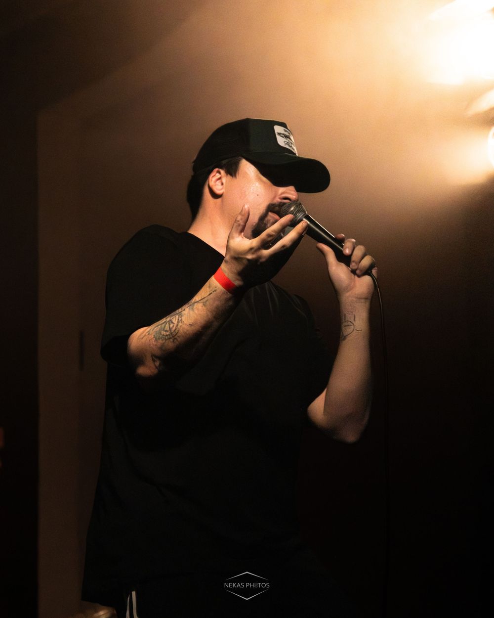 Tattooed male rapper performing on stage holding microphone, wearing black cap and shirt under dramatic spotlight.