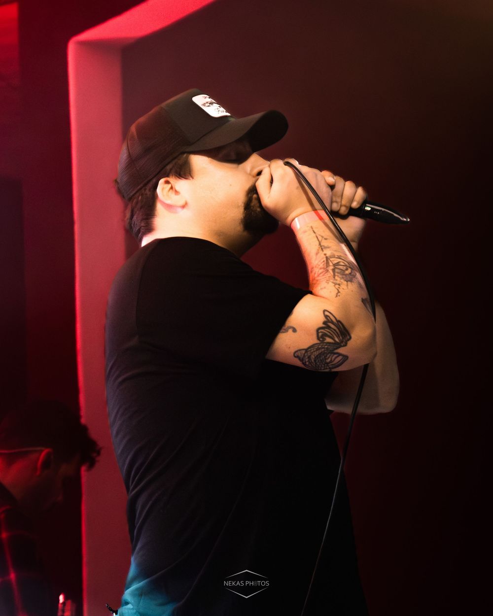 Male rapper performing on stage with microphone, black cap, tattoos on arm, dramatic red lighting.