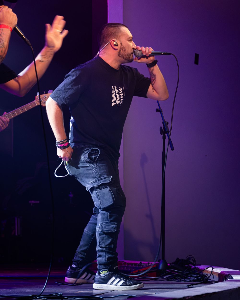 Male vocalist performing on stage, singing into microphone, wearing black t-shirt and cargo jeans, with guitarist visible behind him.