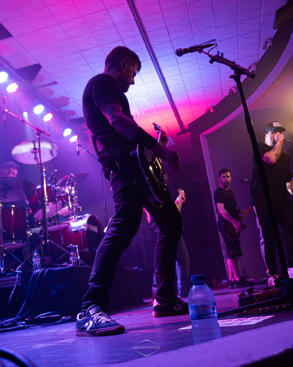 Rock band performing live on stage under purple and pink lights, guitarist in black playing electric guitar.