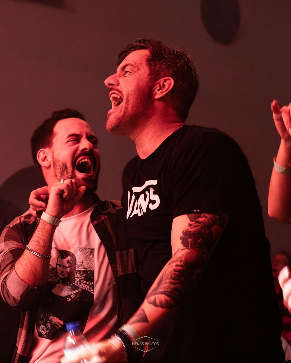 Two tattooed men laughing joyfully at a live music concert under red stage lighting.
